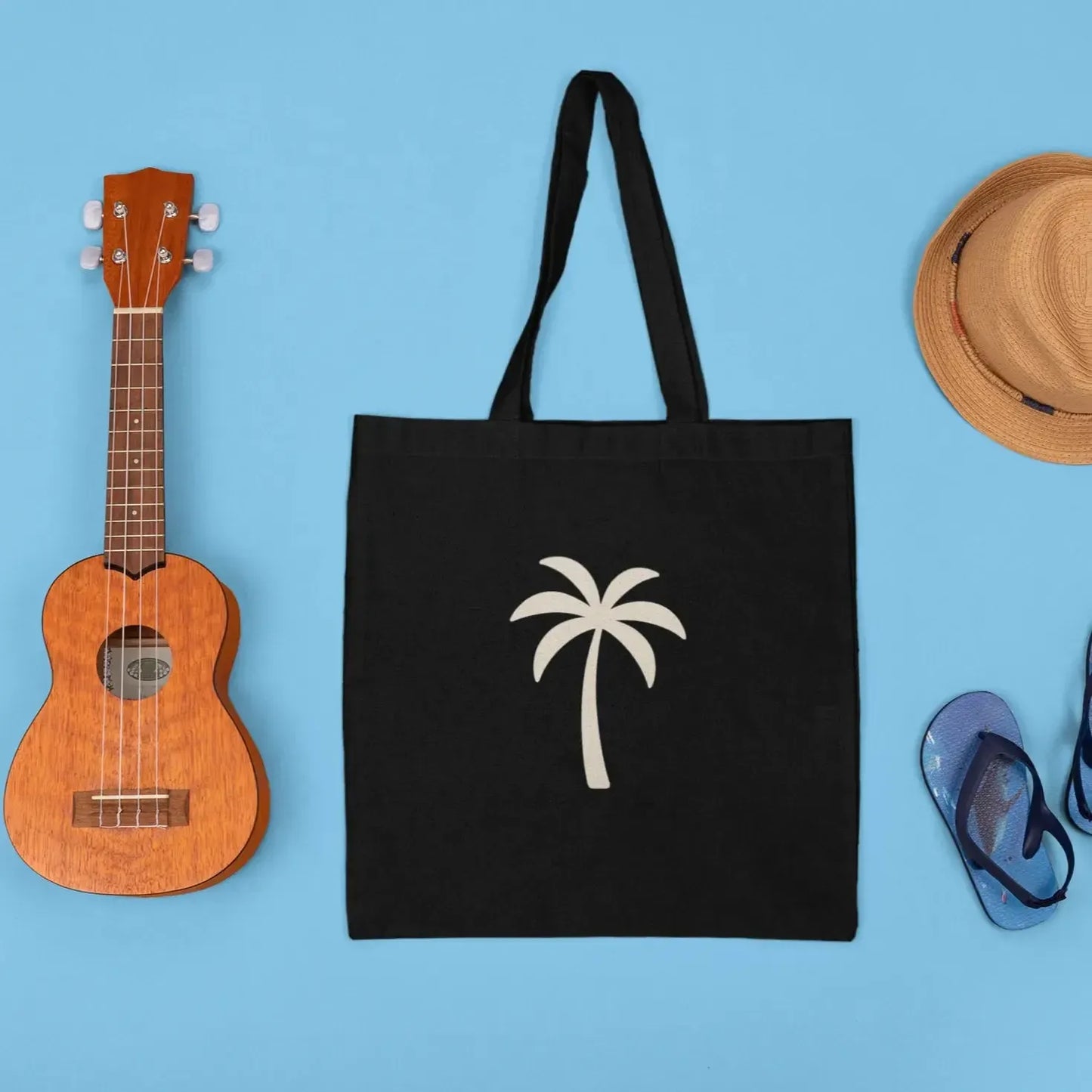 Black tote bag with palm tree design, ukulele, straw hat, and sandals on a blue background